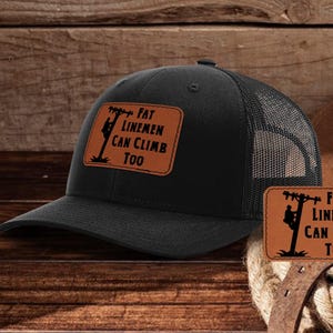 Può includere: Cappellino da camionista nero con una toppa in pelle marrone che dice "Fat Linemen Can Climb Too". La toppa presenta la silhouette di un lineman che scala un palo della luce.