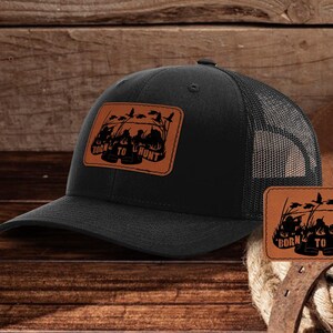 Może przedstawiać: Czarna i brązowa czapka trucker z brązową naszywką z napisem "Born to Hunt" i sylwetką myśliwych z kaczkami.