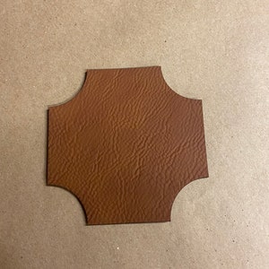 May include: A brown leather coaster with a unique, four-lobed shape. The coaster has a textured surface and a dark brown edge. The background is a light brown surface.