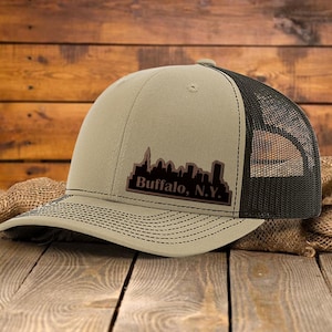 Może przedstawiać: Beżowo-czarna czapka typu trucker z napisem "Buffalo, N.Y." i grafiką panoramy miasta. Czapka ma siatkową tylną część i zakrzywiony daszek z ozdobnymi przeszyciami. Czapka jest prezentowana na drewnianej powierzchni.