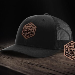 Może przedstawiać: Czarna i brązowa czapka trucker z brązową skórzaną naszywką z napisem "And so the adventure begins" z grafiką gór i drzew.