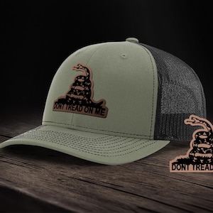 Może przedstawiać: Zielono-czarna czapka trucker z czarno-białym naszywką węża z napisem "Don't Tread On Me". Naszywka znajduje się z przodu czapki, a czapka ma czarny siateczkowy tył.