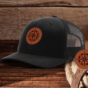 Op de afbeelding: Een zwarte truckercap met een bruine leren kompaspatch op de voorkant. De pet heeft een zwart mesh achterkant.