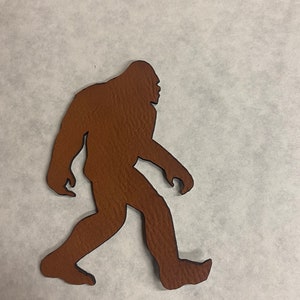 以下が含まれることがあります： 右に向かって歩くビッグフットの茶色のシルエット。シルエットは質感のある素材でできています。