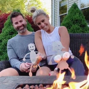 Könnte beinhalten: Zwei Personen rösten Marshmallows über einer Feuerstelle. Die Marshmallows befinden sich auf Metallspießen in Form von Strichmännchen. Die Feuerstelle ist mit Lavasteinen gefüllt.