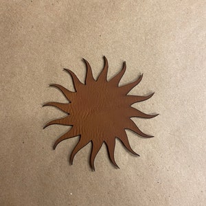 May include: A brown, sun-shaped leather cutout with pointed rays. The cutout is a solid, warm brown colour and is set against a neutral background. The sun design is a decorative element.