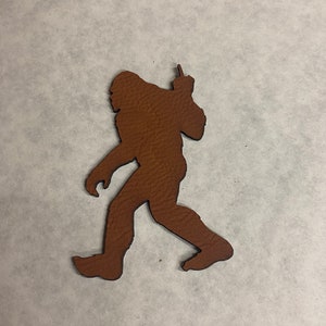 以下が含まれることがあります： 腕を上げて歩いているビッグフットの茶色の革の切り抜き。まるで手を振っているようである。