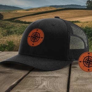 May include: A black trucker hat with a brown leather compass patch on the front. The hat has a black mesh back and a curved brim.