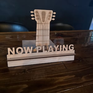 Könnte beinhalten: Ein Schild aus Holz mit der Aufschrift "NOW PLAYING". Das Schild ist an einem Holzausschnitt eines Gitarrenhalses befestigt.