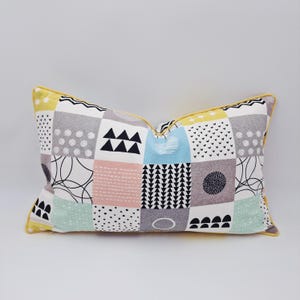 May include: Rectangular decorative pillow with a patchwork design. The pillow features squares in various colors, including gray, yellow, pink, and blue, with geometric and abstract patterns. The pillow has a yellow border.