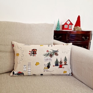May include: A rectangular cushion with a beige background featuring a whimsical print of mice, cheese, and Christmas trees. The cushion is on a beige sofa. A small red house and gnome are on a dark wooden surface.