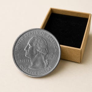 Puede incluir: Una moneda de cuarto de dólar estadounidense en tono plateado con el perfil de George Washington, el texto UNITED STATES OF AMERICA, LIBERTY, IN GOD WE TRUST y la marca de ceca D, junto a una caja cuadrada de cartón abierta con forro negro.
