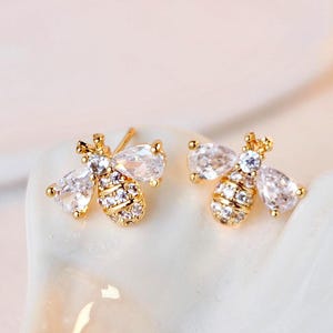 May include: A pair of gold-colored bee-shaped stud earrings. Each earring features a body encrusted with small, clear crystals and pear-shaped crystal wings. The earrings are displayed on a white, curved surface.