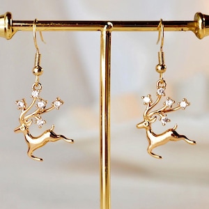 Puede incluir: Un par de pendientes dorados con forma de reno con ganchos colgantes. Cada pendiente presenta una silueta de reno con detalles de cristal en las astas. Los pendientes se exhiben en un soporte para joyas dorado.