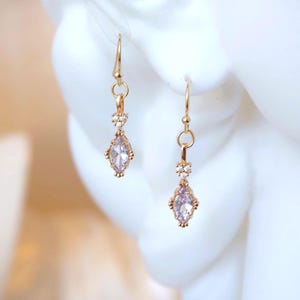 May include: A pair of gold-toned drop earrings featuring a marquise-cut light purple stone at the base, topped with a small cluster of three clear white crystals. The earrings hang from simple gold-toned wire hooks with a delicate metal setting.