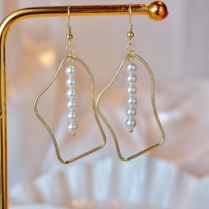 May include: A pair of gold-toned dangle earrings featuring an irregular, open-frame geometric shape. Each earring is suspended from a hook and contains a vertical strand of six small, white, spherical faux pearls centered within the frame.