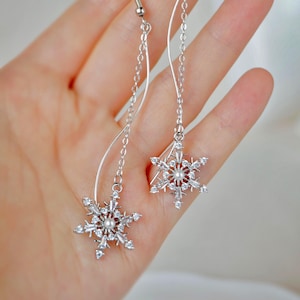 May include: Silver snowflake earrings with clear crystals and a pearl center. The earrings have a long chain and a curved silver wire design. The snowflake design is a winter theme.