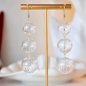 May include: Clear, ribbed glass ball earrings with gold-colored hooks and connecting wires. The earrings are displayed on a gold-colored earring stand. The background is blurred, with hints of white and red.