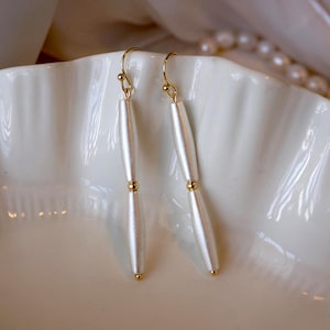 Op de afbeelding: Een paar elegante oorhangers. Elke oorbel heeft een langwerpige, parelmoerwitte kraal, opgehangen aan een goudkleurige haak. Een kleine gouden kraal accentueert het midden van elke oorbel. De oorbellen liggen op een witte, geschulpte schaal.