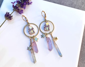 May include: A pair of celestial earrings. Each earring has a gold-tone hoop with a shimmering crystal pendant, dangling star charms, and elongated crystal pendants in purple and clear. A sprig of lavender is in the background.