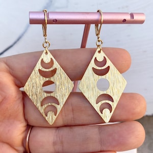 May include: Gold-colored diamond-shaped earrings with a textured finish. Each earring features a design of crescent moons and a central circle cutout. The earrings are displayed on a pink stand.
