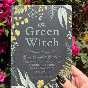 May include: A hardcover book titled The Green Witch: Your Complete Guide to the Natural Magic of Herbs, Flowers, Essential Oils, and More by Arin Murphy-Hiscock, featuring a dark gray cover with yellow and light green botanical illustrations.