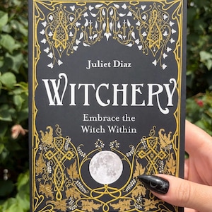 May include: A black book titled Witchery: Embrace the Witch Within by Juliet Diaz. The cover features gold filigree, white leaf illustrations, and a central full moon graphic, all framed by an intricate gold border on a dark background.