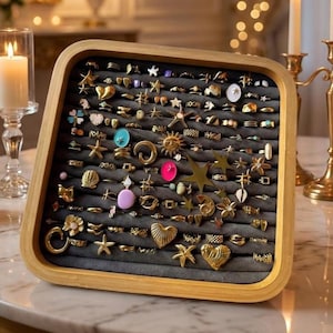 Puede incluir: Una bandeja de exhibición de anillos de madera llena de varios anillos dorados. Los anillos presentan diversos diseños, incluyendo estrellas, corazones y motivos florales. La bandeja está forrada con tela gris y se encuentra sobre una superficie de mármol.