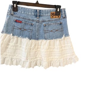 May include: Light blue denim skirt with a white eyelet lace ruffle. The skirt has a classic five-pocket design with the brand name "Mud" on a leather patch. The skirt is hanging on a black hanger.