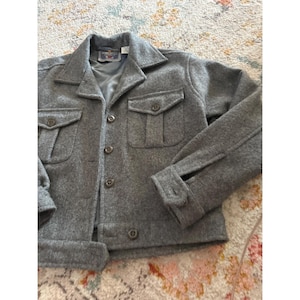 May include: A gray wool-blend jacket featuring a collared neckline, a button-down front, and two chest pockets with buttoned flaps. The long sleeves have buttoned cuffs, and the interior is lined with a smooth, dark gray fabric.