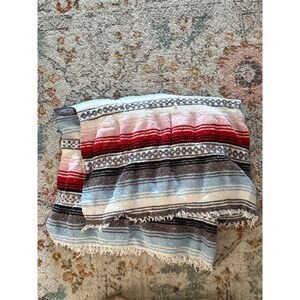 May include: A folded woven blanket featuring horizontal stripes in shades of red, white, light blue, and dark gray. The textile displays a repeating geometric pattern across the center and includes a frayed, fringed edge along the bottom.