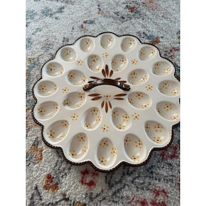 May include: A round ceramic egg serving platter with a scalloped edge and a central brown handle. The white surface features twenty-four egg-shaped indentations, each decorated with small yellow and red painted dots, and a brown leaf motif.
