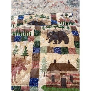 May include: A quilted fabric featuring a patchwork design with illustrations of black bears, a cabin, pine trees, and a moose. The pattern includes squares of plaid, polka dots, and solid colors in shades of brown, green, blue, and cream.