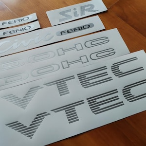 Puede incluir: Un conjunto de calcomanías de coche plateadas con las palabras "Civic", "Ferio", "SiR", "DOHC" y "VTEC".