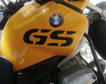 Könnte beinhalten: Nahaufnahme eines gelben Motorrad-Benzintanks mit einem schwarzen "GS"-Logo und einem blau-weißen BMW-Logo.