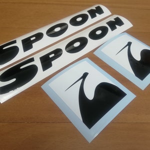Könnte beinhalten: Zwei weiße Aufkleber mit schwarzem Text "SPOON" und zwei weiße Aufkleber mit schwarzen Silhouetten eines Krans.