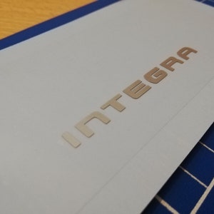 Puede incluir: Un emblema metálico plateado con la palabra "INTEGRA" en letras en relieve.