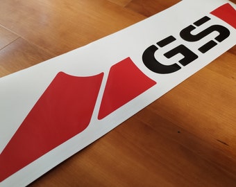 Könnte beinhalten: Weißer Aufkleber mit den schwarzen Buchstaben "GS" und roten Akzentformen. Der Aufkleber befindet sich auf einer Holzoberfläche. Die Buchstaben sind in einer fetten, serifenlosen Schriftart. Die roten Formen sind geometrisch und eckig.