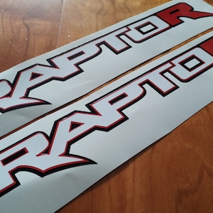 Könnte beinhalten: Zwei weiße Vinyl-Aufkleber mit dem Wort "RAPTOR" in schwarzer und roter Schrift. Die Aufkleber haben einen schwarzen Umriss und einen roten Innenumriss.