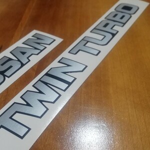 Pode incluir: Dois adesivos prateados em fundos brancos. O primeiro adesivo diz "NISSAN" e o segundo adesivo diz "TWIN TURBO".