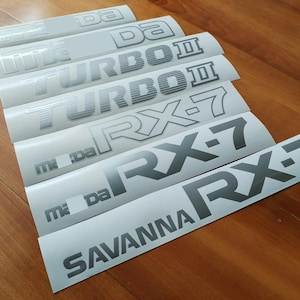 Puede incluir: Un conjunto de ocho calcomanías de vinilo plateadas con varios nombres de modelos de automóviles, que incluyen "RX-7", "TURBO II", "mi da" y "SAVANNA RX-7".