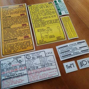 Könnte beinhalten: Eine Sammlung gelber und weißer Warnhinweise für ein Auto. Die Aufkleber enthalten Text in Englisch, Deutsch, Niederländisch, Französisch und Spanisch. Der Text warnt vor dem SRS-System des Autos und den Gefahren, daran zu arbeiten. Die Aufkleber enthalten auch Informationen über die Reifengröße und den Reifendruck des Autos.