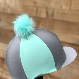 Puede incluir: Un gorro de montar a caballo gris y verde menta con un pompón de color verde menta esponjoso en la parte superior.