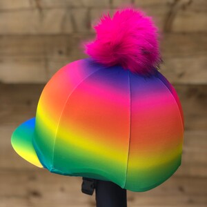 May include: A rainbow ombre riding helmet cover with a pink faux fur pom pom on top. The cover is made of a stretchy, water-resistant material and is designed to fit most riding helmets.