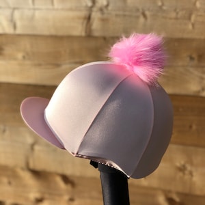 Può includere: Un cappello da equitazione rosa con un pompon in pelliccia sintetica rosa in cima.