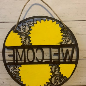 May include: A round wooden welcome sign with a rope hanger. The sign is painted brown with yellow sunflowers and the word "WELCOME" in black lettering.