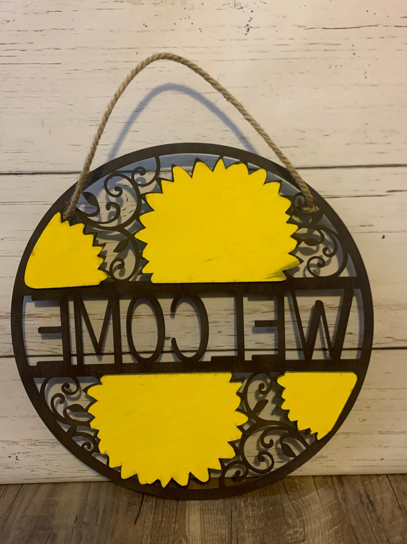 May include: A round wooden welcome sign with a rope hanger. The sign is painted brown with yellow sunflowers and the word "WELCOME" in black lettering.