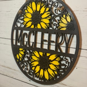 May include: A round wooden sign with a sunflower design and the name "MCCLEERY" in the center. The sign is painted in shades of brown and yellow.