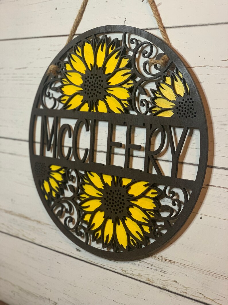 May include: A round wooden sign with a sunflower design and the name "MCCLEERY" in the center. The sign is painted in shades of brown and yellow.