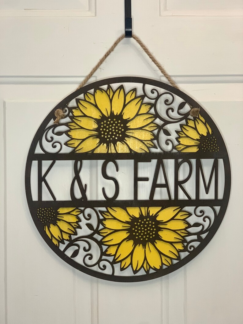 May include: A round wooden sign with a brown background and yellow sunflowers. The sign says "K & S FARM" in black letters.